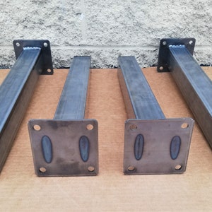 May include: Four metal table legs with square bases and a rectangular plate at the top. The legs are a dark gray color and have a brushed finish.