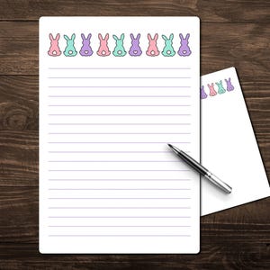 May include: White lined stationery with a row of pastel bunny illustrations across the top. A smaller notepad and a pen are also on the wooden surface. The bunnies are pink, blue, and purple.