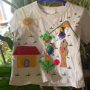 Vintage t-shirt!Village mountain scene with people working, trees, flowers, house ,sun & a mountain on white newborn sized t shirt