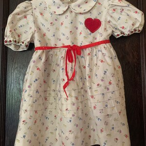 May include: A white dress with a red heart applique and a red ribbon around the waist. The dress has a floral print with small pink, blue, and yellow flowers.