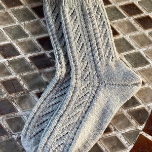 May include: A pair of light gray knitted socks with a diamond pattern. The socks are made of a soft, textured yarn and have a ribbed cuff. The diamond pattern runs down the front of the socks.