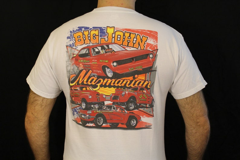 May include: White t-shirt featuring a colorful graphic of vintage red race cars with the text "BIG JOHN Mazmanian". The design includes an American flag motif and various car models, perfect for car enthusiasts.