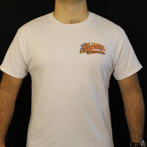May include: White t-shirt with a small, colorful logo on the upper left chest. The logo reads "Big John Magmanian" in a stylized font, with red, white, and blue accents. The shirt is short-sleeved and appears to be made of cotton.
