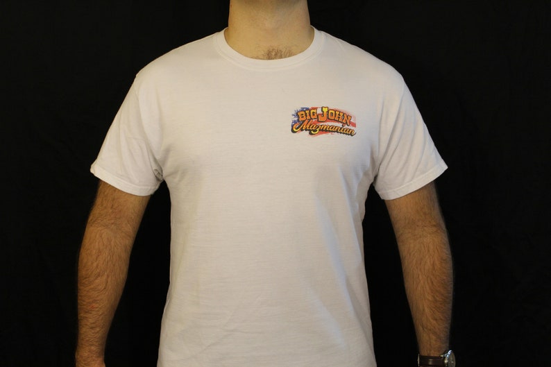 May include: White t-shirt with a small, colorful logo on the upper left chest. The logo reads "Big John Magmanian" in a stylized font, with red, white, and blue accents. The shirt is short-sleeved and appears to be made of cotton.