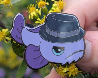 Puede incluir: Un pin de esmalte de pez morado con un sombrero fedora gris oscuro. El pez tiene una expresión de mal humor y un ojo azul claro. El pin está sobre un fondo de flores amarillas y follaje verde.