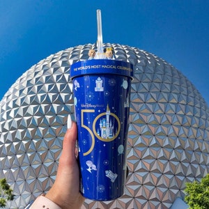 K&ouml;nnte beinhalten: Ein wiederverwendbarer Becher in Blau und Gold mit dem Aufdruck "The World's Most Magical Celebration" und "Walt Disney World 50". Der Becher hat einen Strohhalm und steht vor einer gro&szlig;en, silbernen geod&auml;tischen Kuppel.