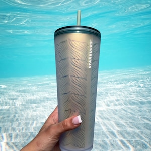 May include: A stainless steel Starbucks tumbler with a white and gold feather pattern. The tumbler has a clear lid and a straw.