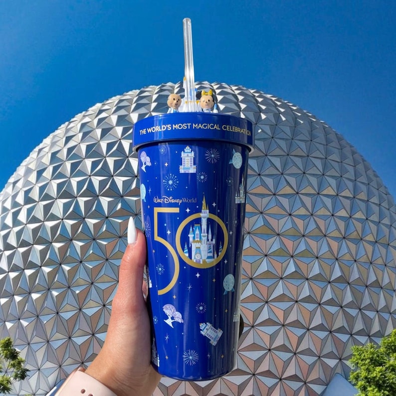 K&ouml;nnte beinhalten: Ein wiederverwendbarer Becher in Blau und Gold mit dem Aufdruck "The World's Most Magical Celebration" und "Walt Disney World 50". Der Becher hat einen Strohhalm und steht vor einer gro&szlig;en, silbernen geod&auml;tischen Kuppel.