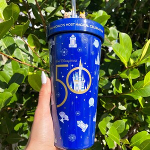 K&ouml;nnte beinhalten: Ein blauer Becher mit einem goldenen 50. Jubil&auml;umslogo und einer wei&szlig;en Silhouette von Schloss Aschenputtel. Der Becher hat einen Strohhalm und ist mit einem blauen Sternenhimmelmuster verziert. Der Text "Walt Disney World" und "The World's Most Magical Celebration" ist auf dem Becher aufgedruckt.