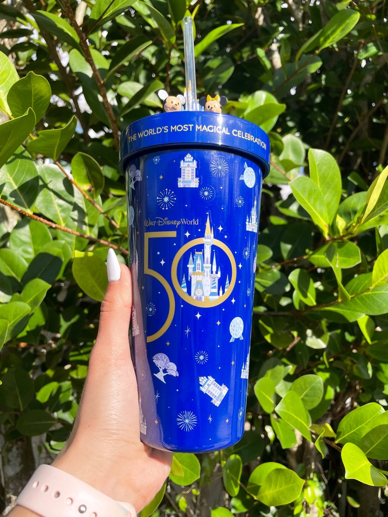K&ouml;nnte beinhalten: Ein blauer Becher mit einem goldenen 50. Jubil&auml;umslogo und einer wei&szlig;en Silhouette von Schloss Aschenputtel. Der Becher hat einen Strohhalm und ist mit einem blauen Sternenhimmelmuster verziert. Der Text "Walt Disney World" und "The World's Most Magical Celebration" ist auf dem Becher aufgedruckt.