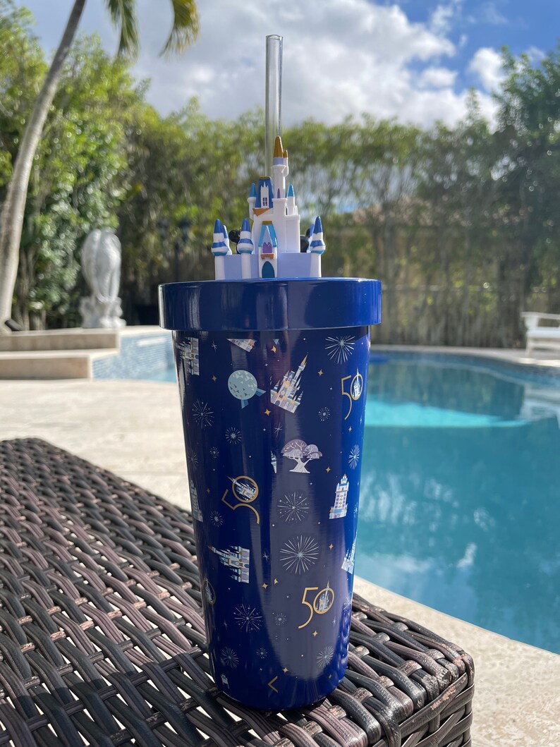 K&ouml;nnte beinhalten: Ein blauer Becher mit einem wei&szlig;en, schlossf&ouml;rmigen Deckel und einem Strohhalm. Der Becher hat ein Muster aus Feuerwerk, Schl&ouml;ssern und der Zahl 50.
