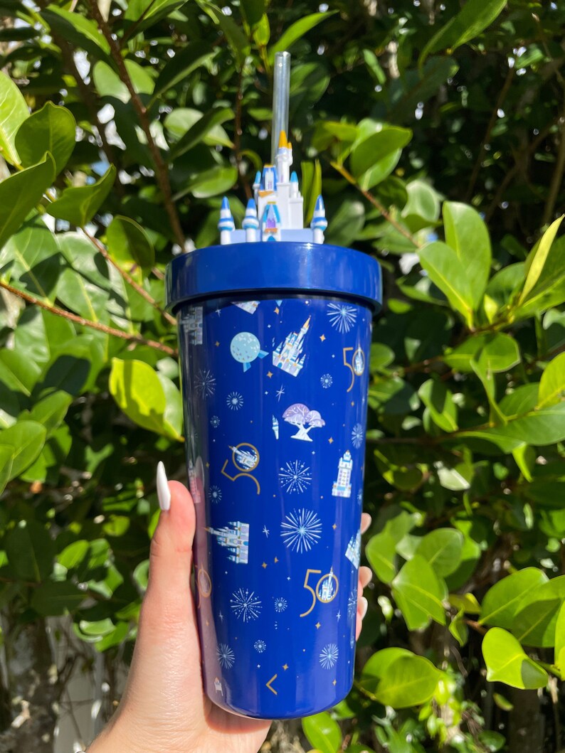 K&ouml;nnte beinhalten: Ein blauer wiederverwendbarer Becher mit einem wei&szlig;en Schlossdeckel und einem Strohhalm. Der Becher hat ein Muster aus wei&szlig;en Schl&ouml;ssern, Feuerwerk und der Zahl 50.
