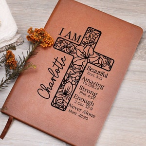 May include: A brown leather journal with the name "Charlotte" and the words "I AM" and inspirational phrases printed on it. A floral cross design is featured prominently. The journal has a ribbon bookmark.