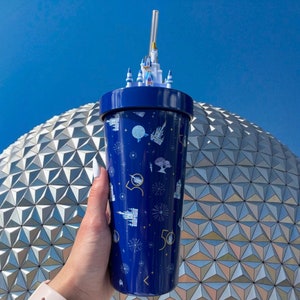 K&ouml;nnte beinhalten: Ein blauer und goldener Becher mit einem 50-j&auml;hrigen Jubil&auml;umsdesign mit dem Schloss von Aschenputtel und Feuerwerk. Der Becher hat einen Strohhalm und einen Deckel. Der Becher steht vor einer wei&szlig;en und silbernen geod&auml;tischen Kuppel.