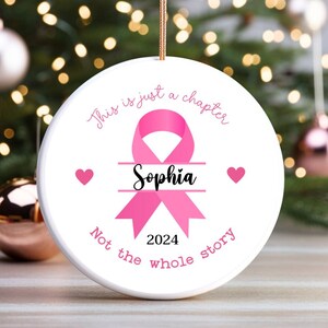 May include: White ceramic Christmas ornament with a pink ribbon design and the name "Sophia". The ornament has the text "This is just a chapter, Not the whole story, 2024" in pink script. Two pink hearts are on either side of the ribbon.