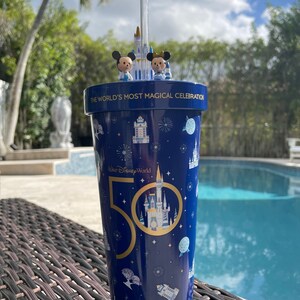 K&ouml;nnte beinhalten: Ein blauer und goldener Becher mit Strohhalm, obenauf Figuren von Mickey Mouse und Minnie Mouse. Der Becher hat ein Design von Schloss Aschenputtel und den Text "The World's Most Magical Celebration" und "Walt Disney World 50".