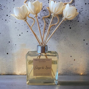 May include: A clear glass bottle with a silver lid and a label that reads "Fidalgo Island Candle Company Reed Diffuser Ginger & Spice 5 Oz. Handmade in Anacortes WA Keep out of reach of children & pets". The bottle is filled with a light yellow liquid and has wooden sticks with white flower tops sticking out of the top.