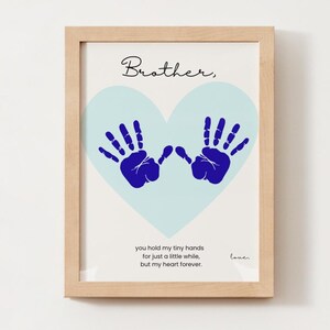Puede incluir: Impresión artística enmarcada con un marco de madera clara. La impresión presenta un corazón azul claro con dos huellas de manos azules. La palabra "Brother," está escrita encima del corazón en cursiva. Debajo, una cita sobre las manos.