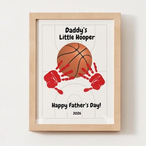 May include: A framed print featuring a basketball court background, a central basketball, and two red handprints. The text reads: Daddy's Little Hooper, Happy Father's Day!, and 2026. The frame is light-colored wood with a clean, simple design.