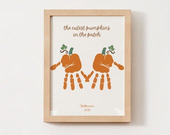 May include: A framed print featuring two orange handprint pumpkins with green stems and leaves. The text reads "the cutest pumpkins in the patch" at the top and "Halloween 2026" at the bottom. The frame is light-coloured wood with a simple design.