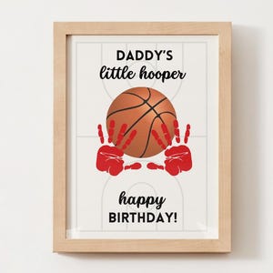 May include: A framed print with a basketball theme. The image features a basketball with red handprints and the text "DADDY'S little hooper happy BIRTHDAY!" in black. The frame is a light wood color.