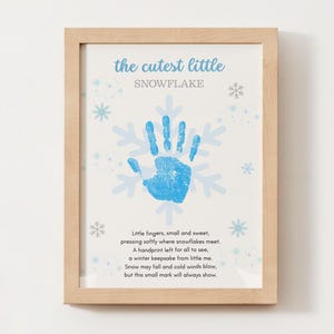 May include: A framed print with a light wood frame. The print features a blue handprint in the shape of a snowflake, with the words "the cutest little snowflake" above it. A poem about a handprint is below the handprint.