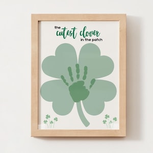 May include: A framed print featuring a large green four-leaf clover with a handprint in the center. The text "the cutest clover in the patch" is written in green script above the clover. Small shamrocks are at the bottom.