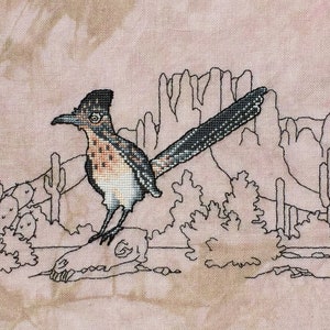 May include: A black and white cross-stitch pattern of a roadrunner bird standing on a skull in a desert landscape with cacti and mountains.