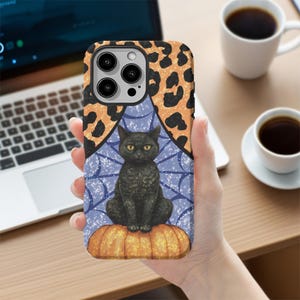 May include: A phone case featuring a black cat sitting on a pumpkin, with a blue spiderweb design and a leopard print border. The case is held in a hand, with a laptop and coffee cups in the background.