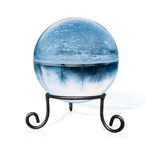 K&ouml;nnte beinhalten: Eine Glaskugel mit einer Winterlandschaft im Inneren, auf einem schwarzen Metallst&auml;nder. Die Szene zeigt eine verschneite Landschaft mit B&auml;umen, die sich im Wasser spiegeln.