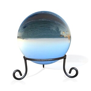K&ouml;nnte beinhalten: Eine Glaskugel mit einem blauen und wei&szlig;en Bild einer Meereslandschaft im Inneren, die auf einem schwarzen Metallst&auml;nder mit drei Beinen steht.