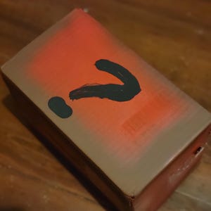 May include: A rectangular cardboard box with a brown base color, featuring a spray-painted orange gradient across the top surface and a large, hand-painted black question mark symbol centered prominently on the lid of the container.