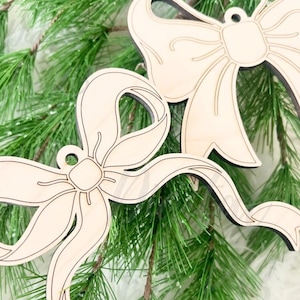 May include: Two wooden bow ornaments with a light beige finish and dark edges are displayed against a backdrop of green pine needles. The ornaments have a detailed ribbon design. The image includes the text "Digital by KB", "SVG", and "Digital Files for Lasers".