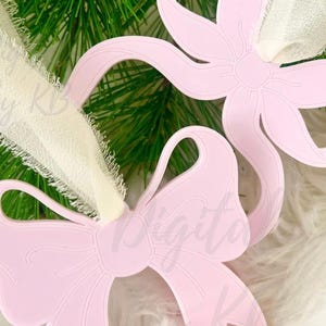 May include: Two light pink bow-shaped ornaments with detailed edges are displayed against a backdrop of greenery and white fabric. The image includes text that reads "Digital by KB" and "Digital Files for Lasers."