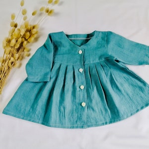 May include: A teal blue linen dress with long sleeves and a button-up front. The dress has a gathered skirt and a simple, classic design.