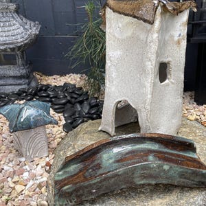 May include: Handmade ceramic garden sculptures, including a white tower with a brown roof, a small bridge, and a miniature pagoda with a blue roof. The sculptures are set on a rock and surrounded by pebbles.