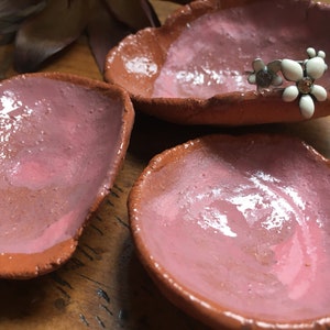 May include: Three small, terracotta-coloured ceramic dishes with a pink glaze. The dishes are slightly irregular in shape and have a rough, textured edge. One dish has a silver and white flower pin on top.