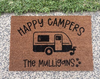 May include: Brown coir doormat featuring the text "HAPPY CAMPERS" above a black campervan illustration. Below the campervan is "The Mulligans" and paw prints. The doormat is rectangular and rests on a concrete surface.