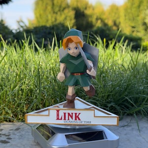 May include: A low-poly Link figurine with orange hair, a green tunic, and brown boots, holding a sword and shield. The figure stands on a triangular white base with gold trim featuring the red text LINK and OCARINA OF TIME, mounted on a silver stand.