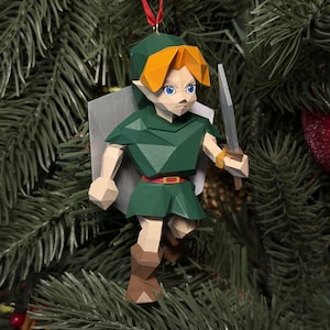 Peut inclure: Une décoration suspendue de style low-poly représentant un personnage aux cheveux blonds, vêtu d'une tunique verte, de bottes marron et d'une ceinture rouge. La figurine tient une épée grise et porte un bouclier gris, suspendue par un ruban rouge.