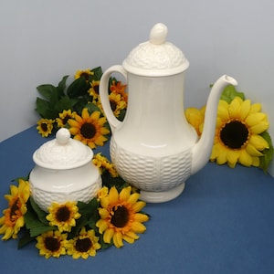May include: White ceramic teapot and sugar bowl with a woven pattern. The teapot has a lid with a knob on top. The sugar bowl has a lid with a knob on top. Both pieces are surrounded by yellow sunflowers.