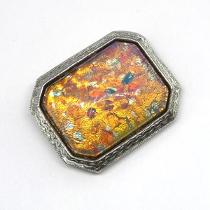 May include: A rectangular brooch with a silver-colored, ornate frame. The center features a vibrant, multi-colored glass design with shades of orange, yellow, and hints of blue and green. The brooch is set against a white background.