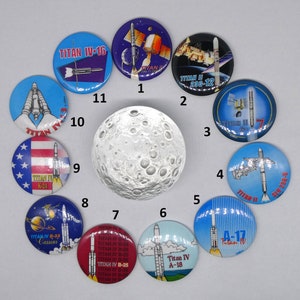 May include: A collection of 12 round, colorful buttons with images of rockets and spacecrafts. The buttons have white text with the words "TITAN IV" and various numbers and letters. The buttons are arranged in a circle around a gray, textured, round object.