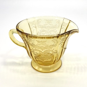 May include: A yellow glass creamer with an embossed floral design. The creamer has a handle and a flared lip.
