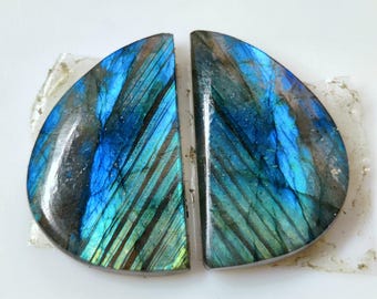 May include: Two semi-circular labradorite gemstones featuring a polished surface with iridescent blue and green flashes. The stones display natural dark inclusions and linear striations across their smooth, reflective, and curved surfaces.