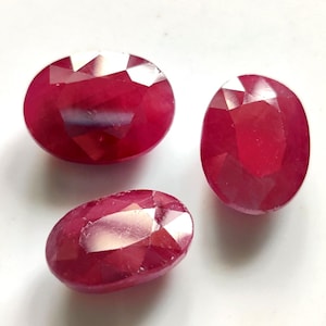 Puede incluir: Tres rubíes rojos facetados de forma ovalada con un acabado pulido. Las piedras preciosas exhiben un tono carmesí profundo y saturado, dispuestas en un grupo que muestra sus superficies reflectantes y sus cortes geométricos precisos.