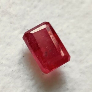 Może przedstawiać: Prostokątny, fasetowany kamień szlachetny rubinu. Klejnot ma głęboki, nasycony czerwony kolor z widocznymi wtrąceniami wewnętrznymi. Kształt to klasyczny szlif szmaragdowy.