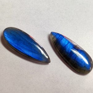 May include: Two polished labradorite gemstones shaped as elongated teardrops. Each stone displays a vibrant, iridescent blue flash across its smooth, curved surface, contrasting with darker, natural mineral inclusions visible at the edges.