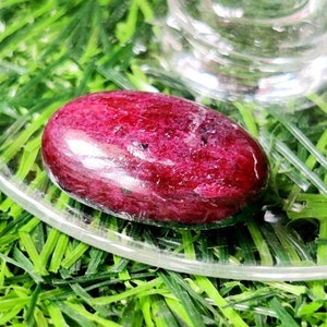 Puede incluir: Una piedra preciosa roja ovalada lisa con un tono rojo ligeramente más oscuro en el centro. La piedra preciosa está sobre una superficie de vidrio transparente.