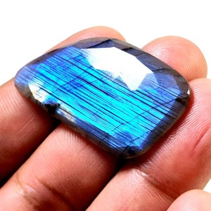 May include: A blue and iridescent labradorite gemstone with a faceted, rectangular shape. The gemstone is held in a hand.